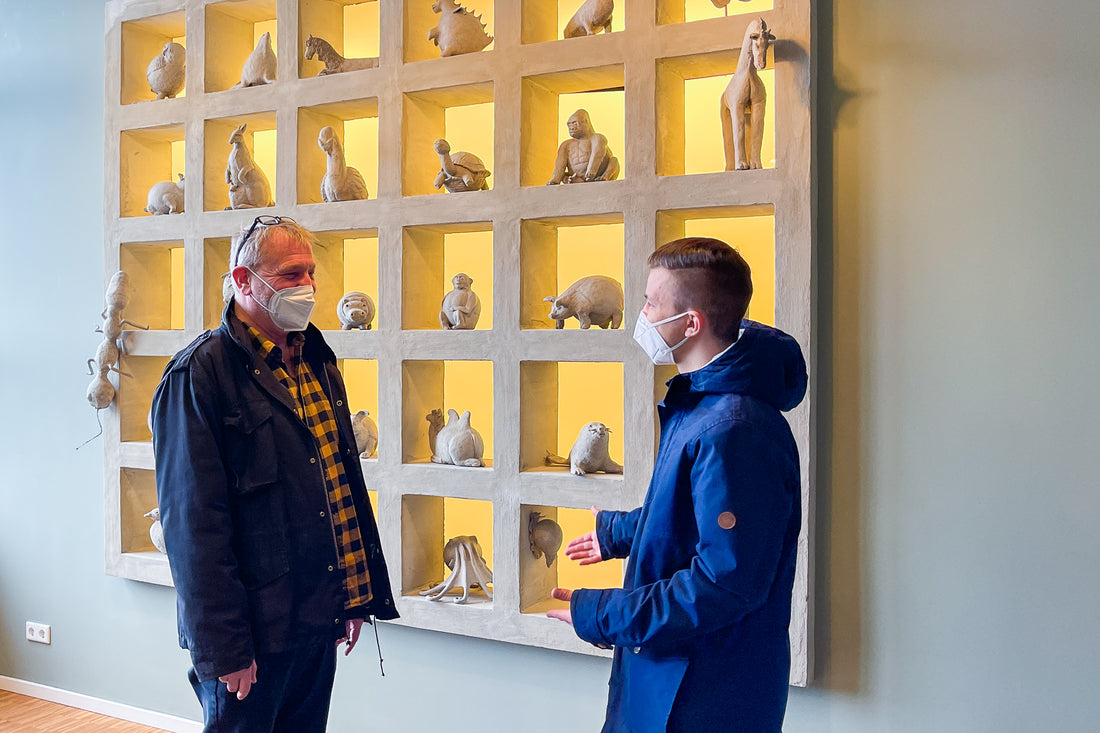 Blumenfisch-Mitarbeiter Ronald Bluhm und Special Olympics Praktikant Julian Stegemann. Im Hintergrund sind Kunstwerke der Mitarbeiter in einem Regal sichtbar.