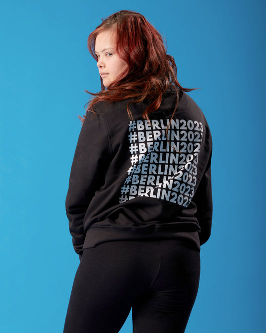 Mädchen steht mit Rücken zur Kamera und schaut in die Kamera. Sie trägt ein schwarzes Sweatshirt mit #Berlin2023 Schriftzug auf dem Rücken