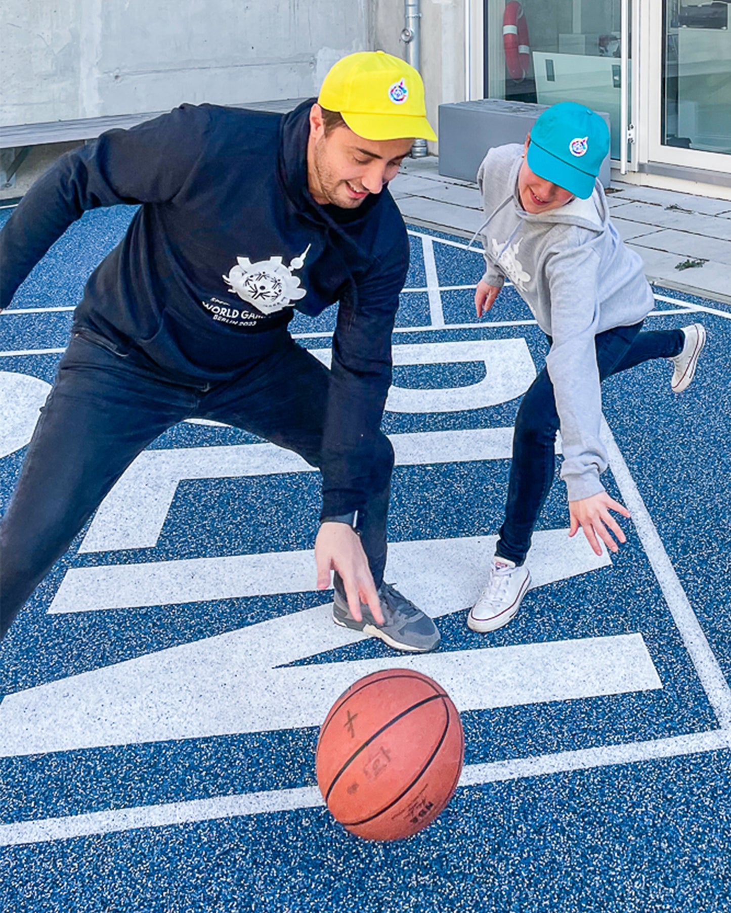 Eine Frau und ein Mann spielen Basketball, der Mann trägt einen schwarzen Hoodie mit Weltspiele Logo und die Frau trägt einen grauen Hoodie mit Weltspiele Logo 