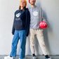 Frau trägt schwarzen Hoodie mit Weltspiele Logo, Mann trägt grauen Hoodie mit Weltspiele Logo und hält einen roten Ball in der Hand