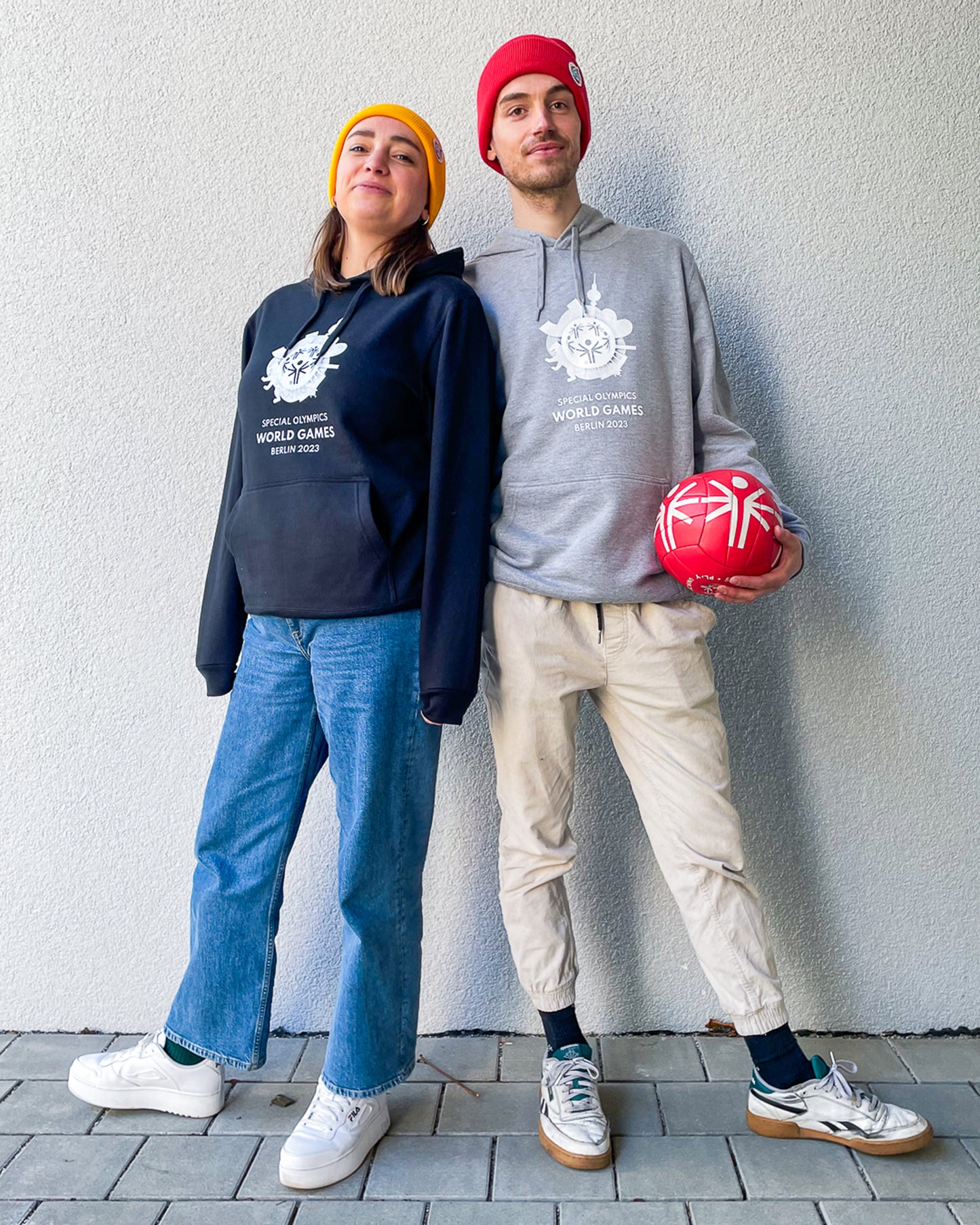 Frau trägt schwarzen Hoodie mit Weltspiele Logo, Mann trägt grauen Hoodie mit Weltspiele Logo und hält einen roten Ball in der Hand
