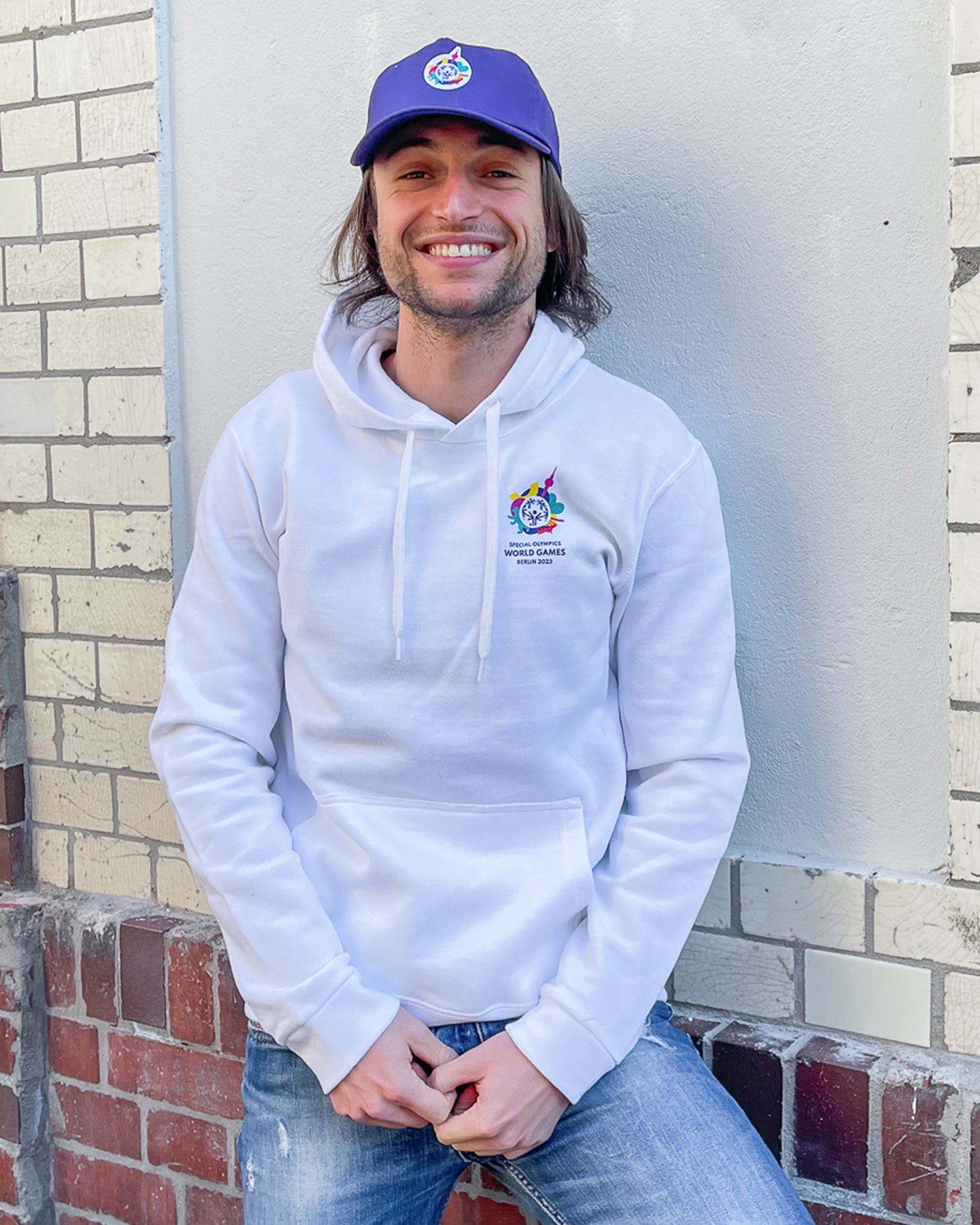 Mann trägt weißen Hoodie mit kleinem Weltspiele Logo auf der Brust und violettes Cap mit Weltspiele Logo 