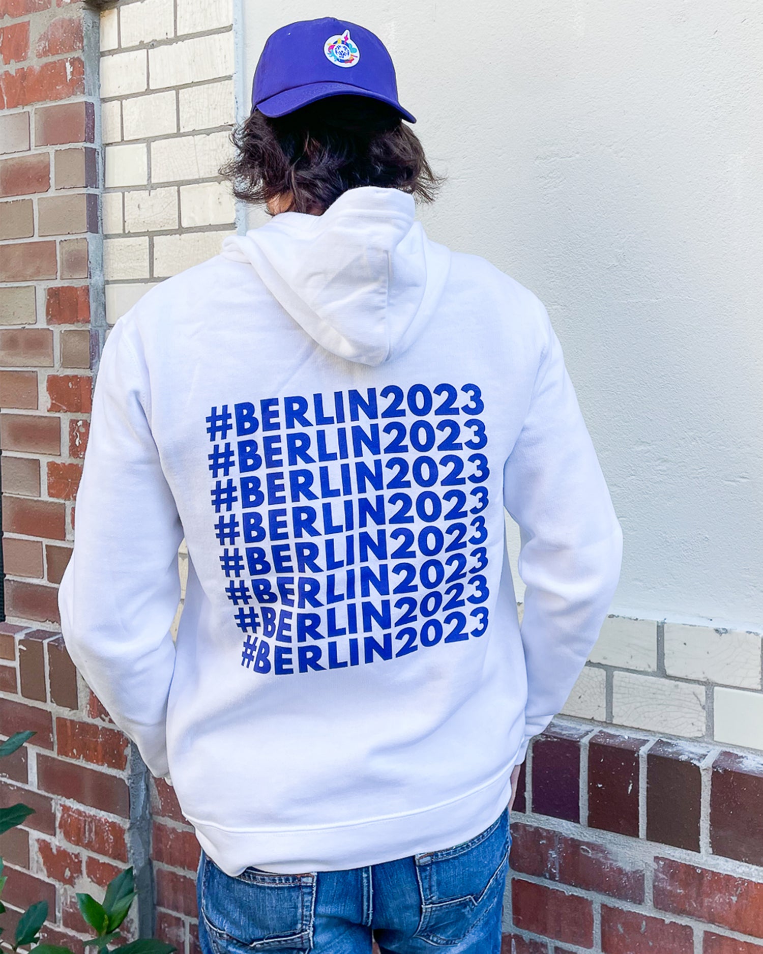 Mann steht vor einer Wand mit dem Rücken zur Kamera. Er trägt weißen Hoodie mit #Berlin2023 Schriftzug auf dem Rücken und violettes Cap mit Logo
