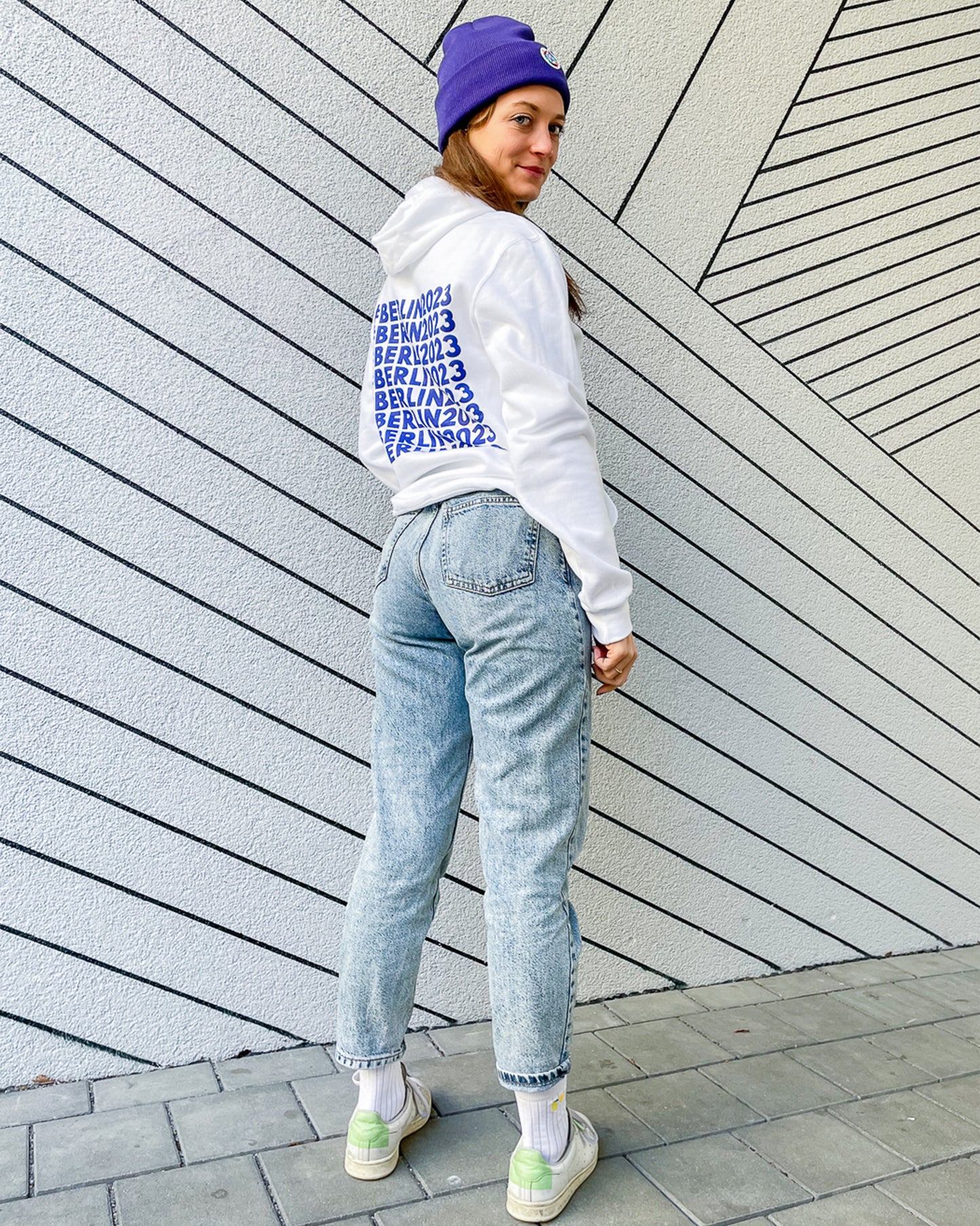 Frau trägt weißen Hoodie mit Weltspiele Logo und violette Mütze mit Logo 