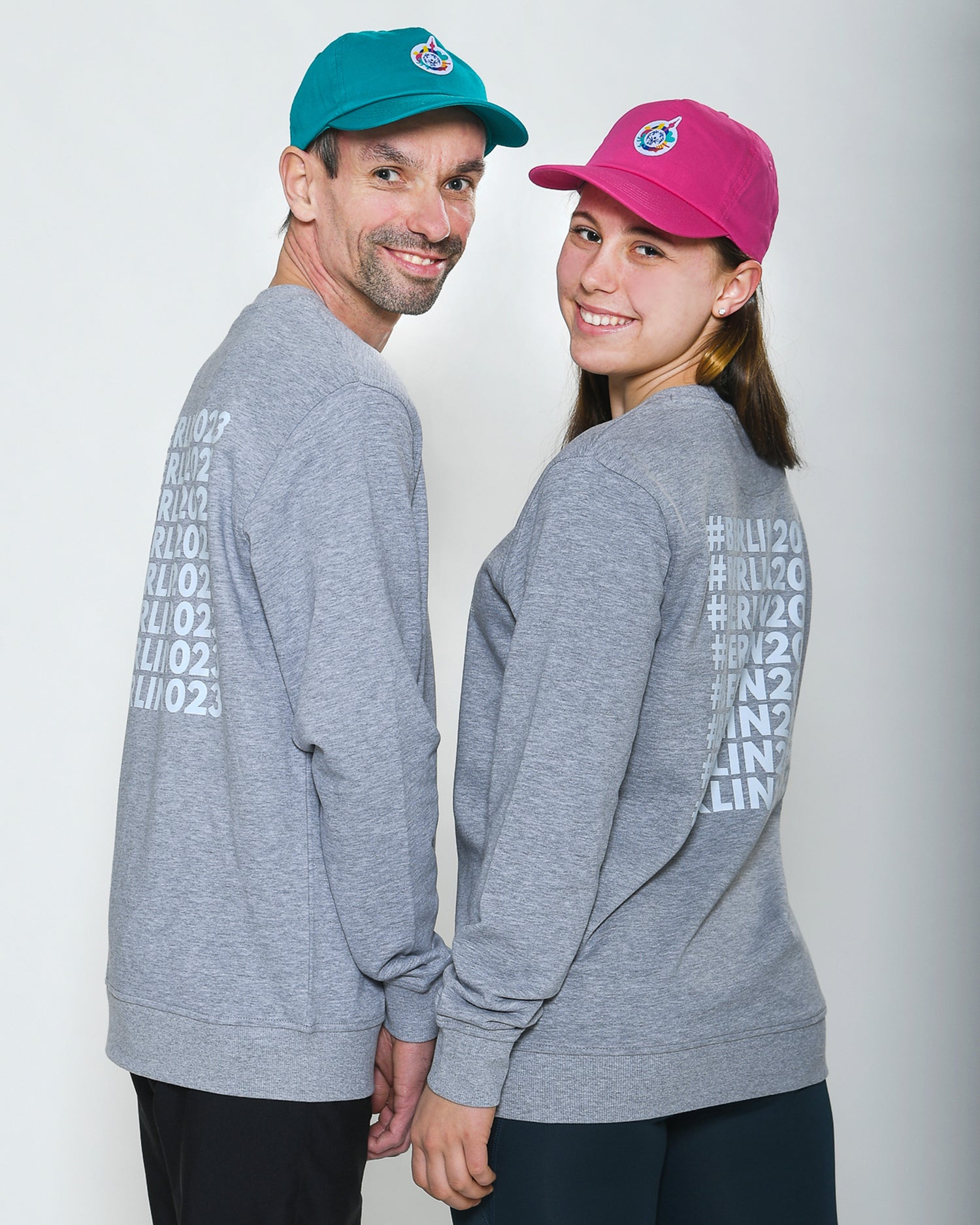 Mann und Mädchen stehen nebeneinander und schauen in die Kamera. Der Mann trägt ein graues Sweatshirt und eine Cap in Petrol mit Logo. Das Mädchen trägt ein graues Sweatshirt und pinke Cap mit Logo. 