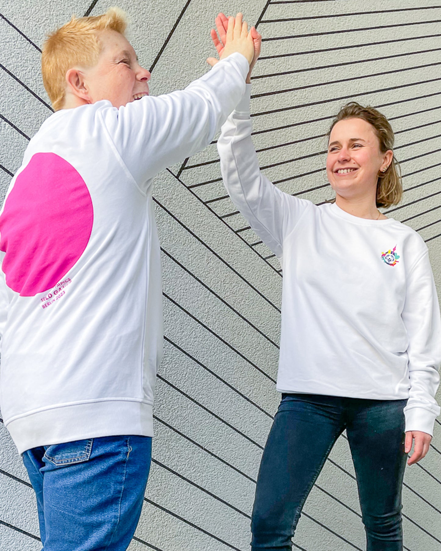 Zwei Frauen klatschen einander ab. Sie tragen ein weißes Sweatshirt mit kleinem Weltspiele Logo auf der Brust und einem großen, pinken Kreis auf dem Rücken.