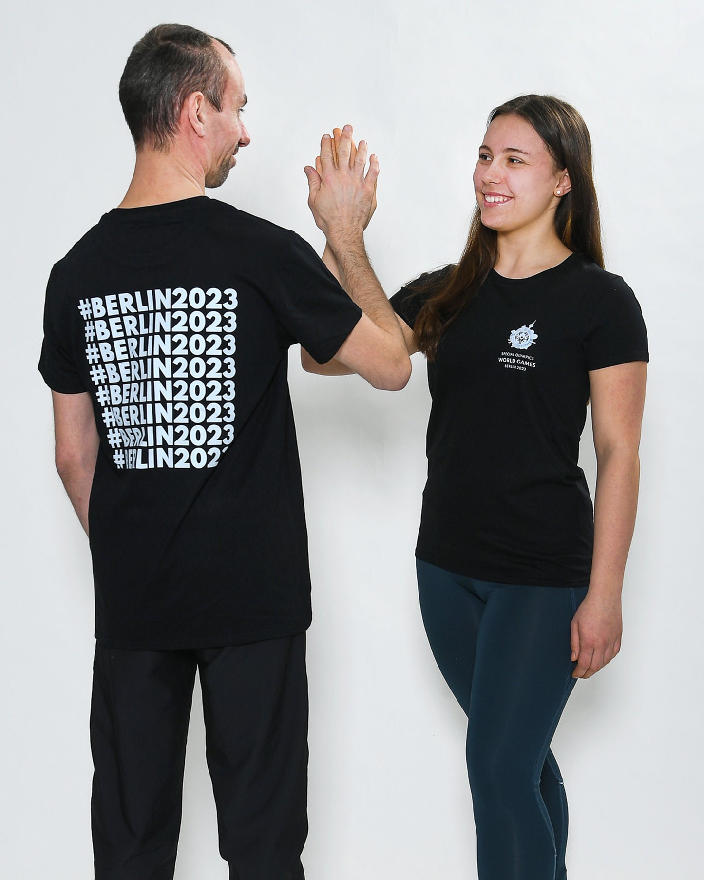 Mann und Frau klatschen einander ab. Sie tragen ein schwarzes Shirt mit einem weißen Weltspiele Logo auf der Brust. Der Mann steht mit dem Rücken zur Kamera, man sieht den #Berlin2023 Schriftzug auf dem Rücken.